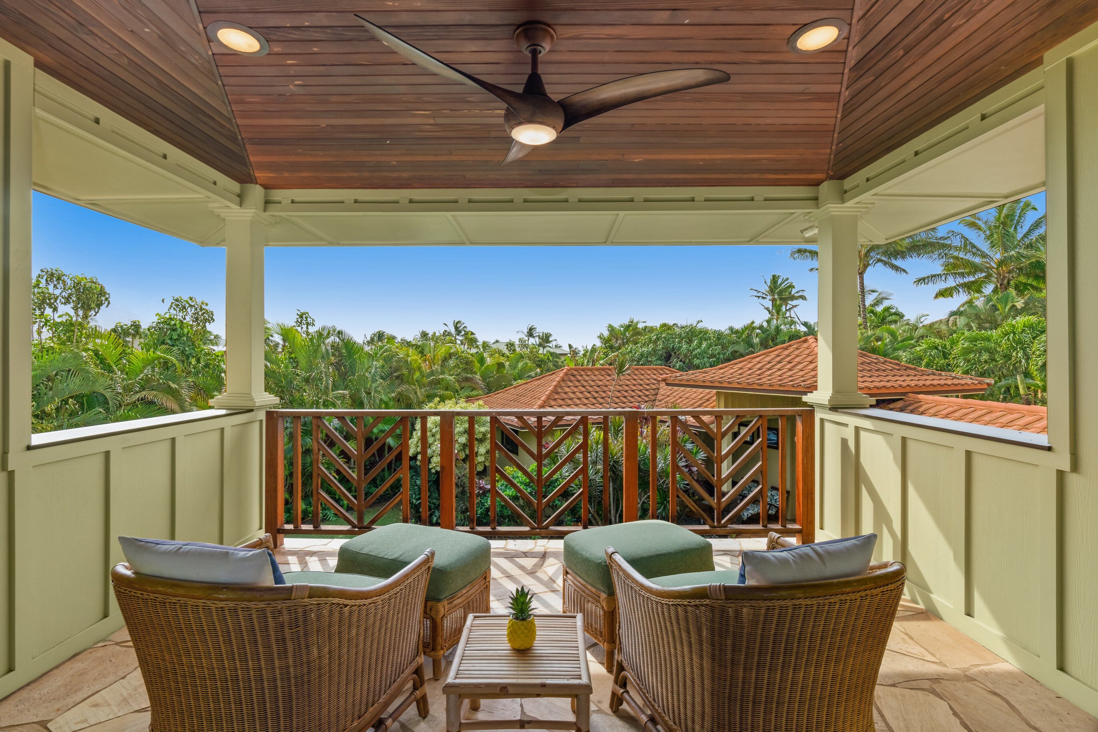 A covered outdoor seating area with wicker furniture and a ceiling fan, overlooking a lush garden and a red-tiled roof building.