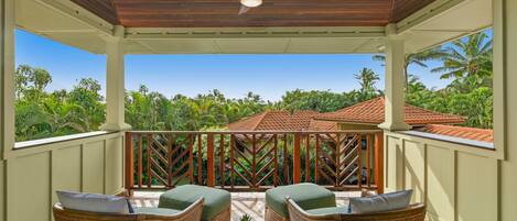 A covered outdoor seating area with wicker furniture and a ceiling fan, overlooking a lush garden and a red-tiled roof building.