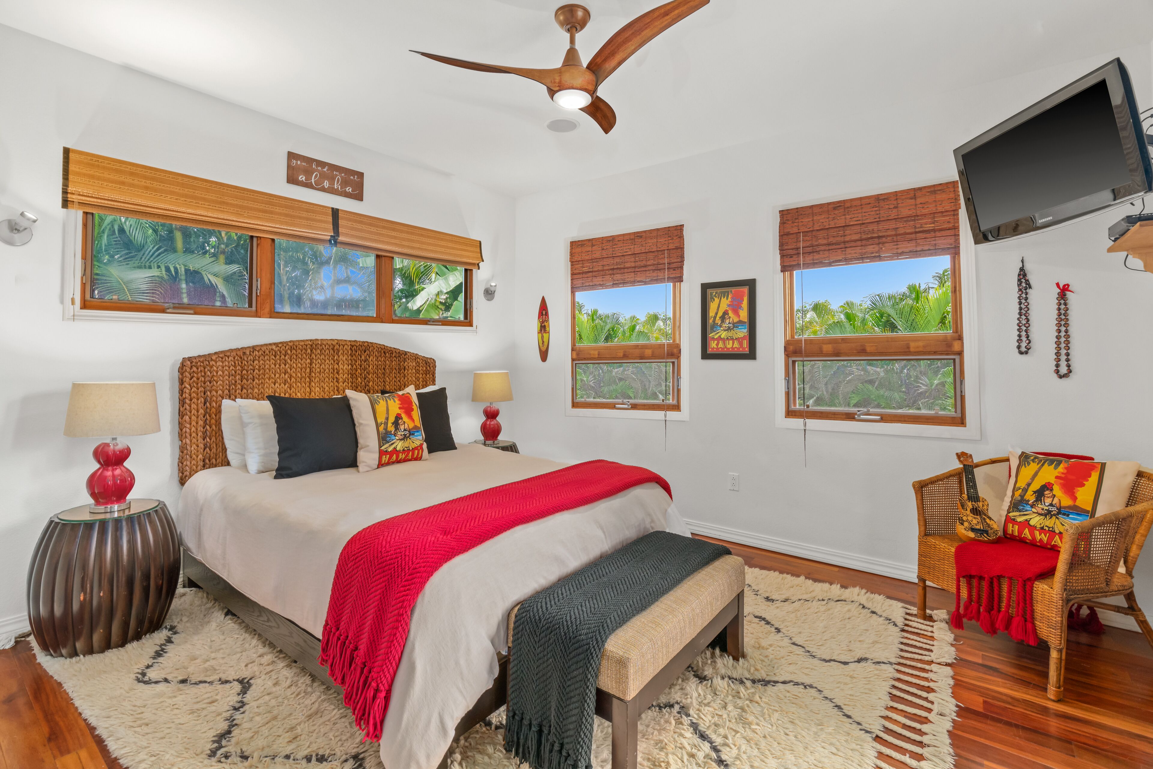 A bedroom with a bed, a ceiling fan, a TV, and large windows with views of greenery.
