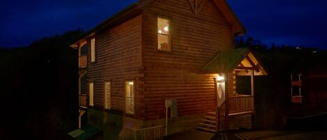 A two-story log cabin with a porch and a small attached structure, surrounded by trees.