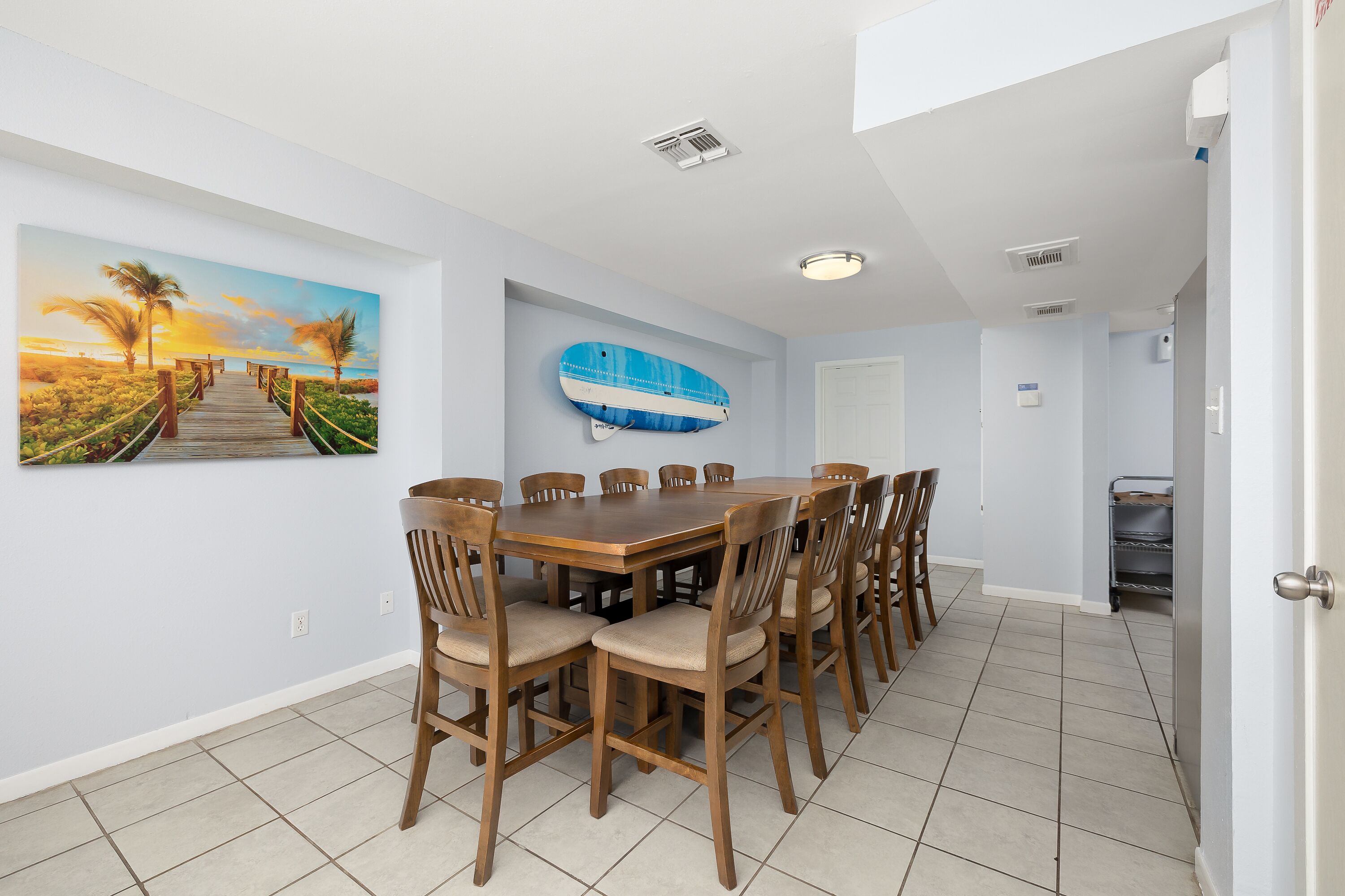 A dining area with a wooden table and chairs, a large painting of a sunset, and a blue surfboard on the wall.