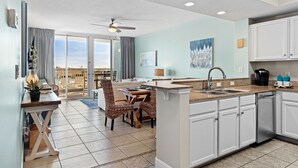 A modern kitchen with white cabinets, a dining area with wicker chairs, and a view through the glass doors. - Waters Edge 109 ~ Ground Floor and Beach Front ~ Steps to Pool and Sand! (Fort Walton Beach)