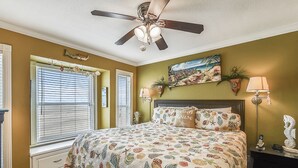 Un dormitorio con cama, ventilador de techo, ventana con persianas, mesita de noche con lámpara y un cuadro en la pared.