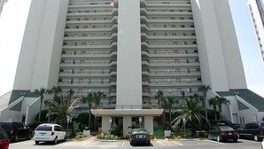 Ein mehrstöckiges Hotel mit zentralem Eingang, Parkplatz und Palmen.