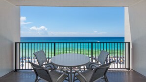 Ein Balkon mit einem runden Tisch und vier Stühlen mit Blick auf einen Strand, auf dem Sonnenschirme und Menschen sind.