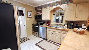 Una cocina compacta equipada con refrigerador, microondas, estufa y lavavajillas. Hay un fregadero con encimera y una cafetera. Los gabinetes son de colores claros y cuenta con un salpicadero decorativo.