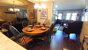 Un comedor con una mesa redonda de madera, sillas metálicas y una araña. Hay cuadros en las paredes y una ventana con cortinas.