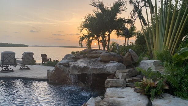 Zona de piscina con cascada, sillas de descanso y vista al atardecer sobre el agua.