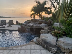 Ein Poolbereich mit Wasserfall, Liegestühlen und Blick auf den Sonnenuntergang über dem Wasser.