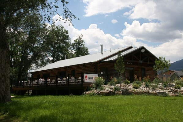 A rustic log cabin with a balcony, surrounded by greenery and mountains in the background.