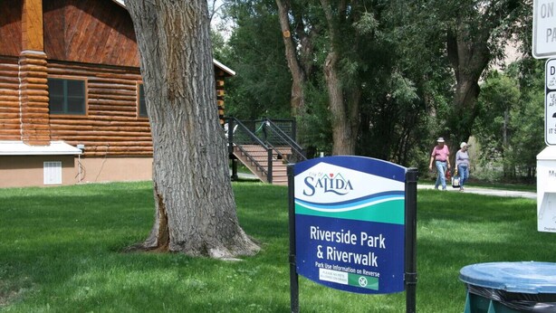 A sign for Riverside Park & Riverwalk in Salida, with a wooden building and a large tree in the background. - Comfort by the river & short walk to downtown. (Salida)