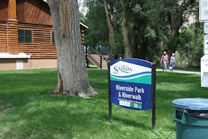 A sign for Riverside Park & Riverwalk in Salida, with a wooden building and a large tree in the background.