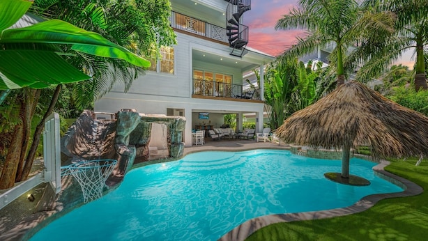 A pool with a waterfall feature, surrounded by lush greenery and a thatched roof structure.