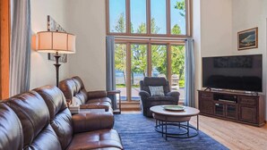 A living room with a leather sofa, a round coffee table, a television on a cabinet, and large windows with a view of trees. - Beside Quiet Waters | Waterfront | Lake Bellaire (Bellaire)