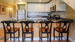 A kitchen with white cabinets, a stainless steel refrigerator, a microwave, and a stove. There are three wooden chairs with black backs. - Keone Nani Condo - New AC, Golf, Swim, Food Trucks (Kahuku)