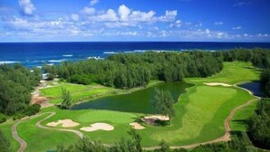 A golf course with a water hazard, surrounded by dense trees and a view of the ocean. - Keone Nani Condo - New AC, Golf, Swim, Food Trucks (Kahuku)