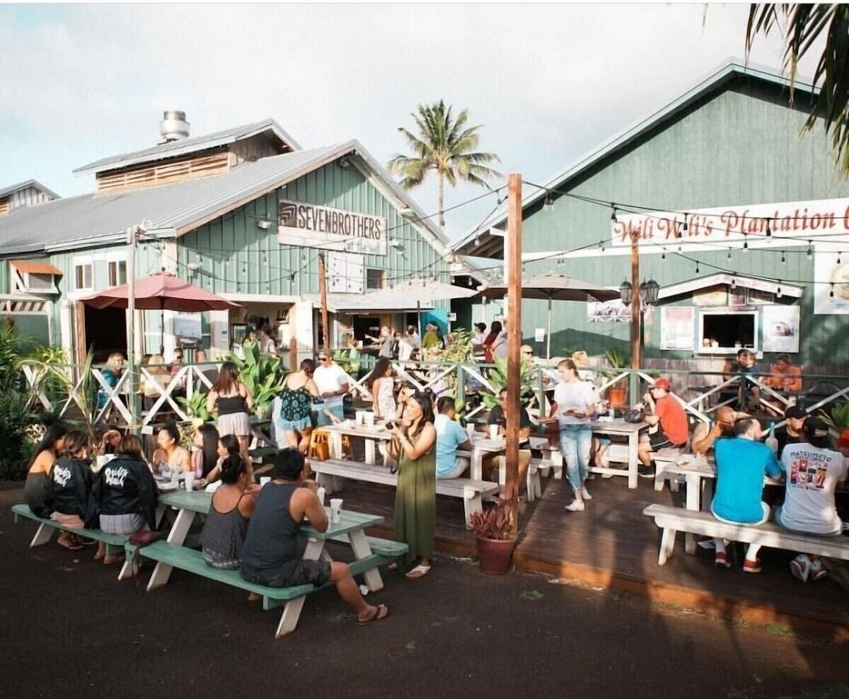 An outdoor dining area with multiple tables and benches, surrounded by buildings with signs, including one that reads "SEVENBROTHERS."