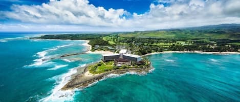 An aerial view of a coastal hotel with a central building surrounded by lush greenery and a clear view of the ocean.