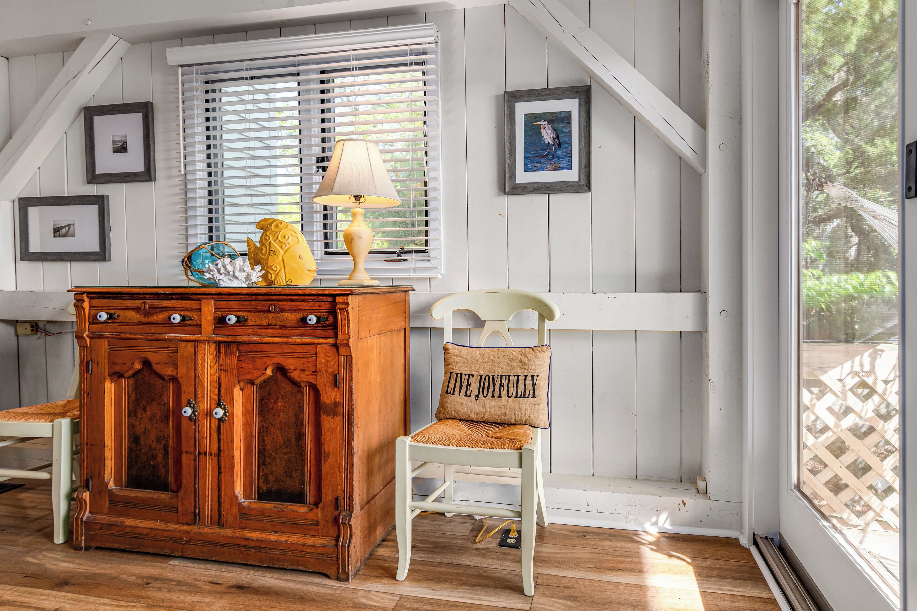 A cozy room with a wooden cabinet, a chair with a cushion, a lamp, and framed pictures on the wall.