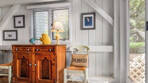 A cozy room with a wooden cabinet, a chair with a cushion, a lamp, and framed pictures on the wall. - Beautiful sunsets at this marsh view condo; enjoy the community pool close to all islands amenities (Bald Head Island)