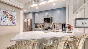 A modern kitchen with light blue cabinets, a white island, wicker chairs, and a herringbone-patterned backsplash. - Beautiful sunsets at this marsh view condo; enjoy the community pool close to all islands amenities (Bald Head Island)