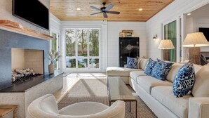 A living room with a fireplace, a ceiling fan, a sofa with patterned cushions, and a large window offering a view of trees. - Magnificent East Beach Home! Heated Pool, 5BR/5.5BA Sleeps 16 (St. Simons Island)