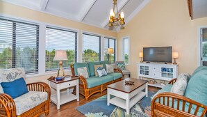 A spacious living room with wicker furniture, a television, and large windows with blinds.