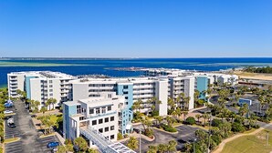 A coastal hotel complex with multiple buildings, parking, and a marina in the background.