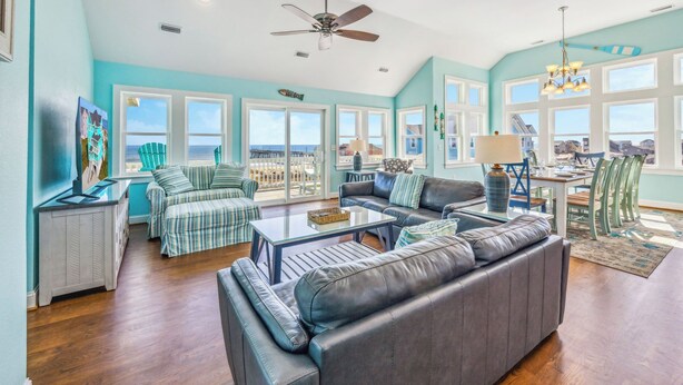 A spacious living room with a large gray sofa, striped armchairs, and a glass coffee table. There are multiple windows offering a view of the ocean. - Premier Oceanfront Dog Friendly w/ Pool, Hot Tub, Elevator, Theater and Game Rms (Rodanthe)