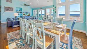 A dining area with a large table set for six, surrounded by white chairs, with a view of the ocean through large windows. - Premier Oceanfront Dog Friendly w/ Pool, Hot Tub, Elevator, Theater and Game Rms (Rodanthe)
