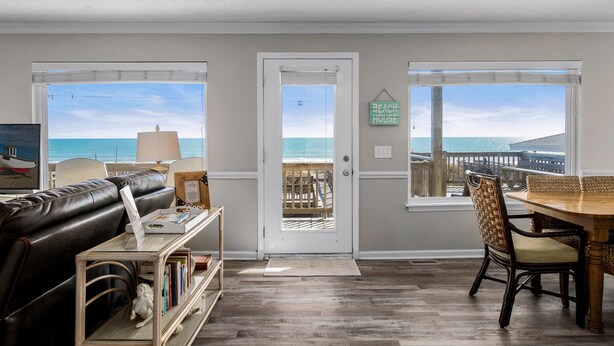 Une pièce avec vue sur l’océan, une porte sur laquelle est apposée une plaque indiquant « Maison de plage », une table à manger avec des chaises, un canapé en cuir et une bibliothèque.