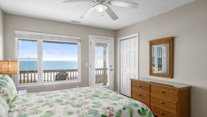 Une chambre à coucher avec un lit, un ventilateur de plafond, une grande fenêtre donnant sur l’océan, une porte et une commode en osier.