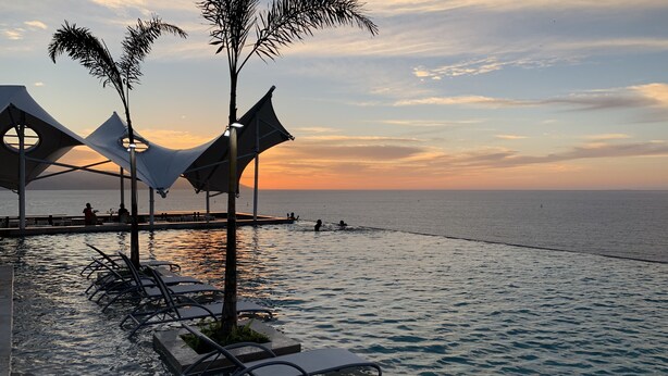 Ein Poolbereich mit Liegestühlen und einer Palme mit Blick auf das Meer bei Sonnenuntergang.