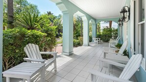 Een overdekt terras met witte Adirondack-stoelen en een betegelde vloer, omgeven door weelderige begroeiing en een heldere blauwe lucht.
