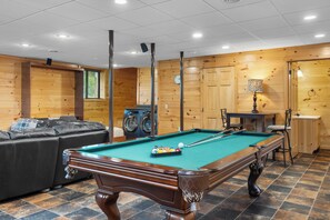 Une table de billard avec des billes, un canapé et un lave-linge dans une pièce aux murs en bois et au sol carrelé.