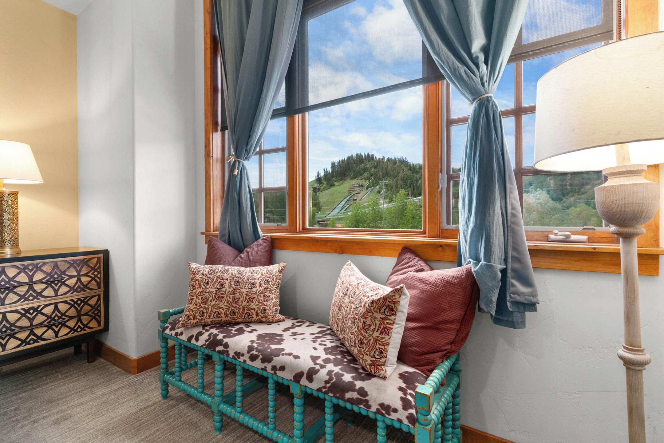 Una habitación acogedora con un banco de madera, cojines estampados, una mesita auxiliar y vistas a un paisaje verde a través de una ventana con cortinas azules.