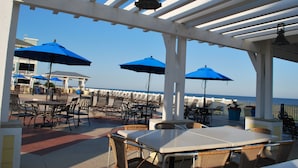 An outdoor patio with tables and chairs, blue umbrellas, and a view of the ocean. - Beachfront at Pointe West. Walk to the beach and the club all just steps away! (Galveston)