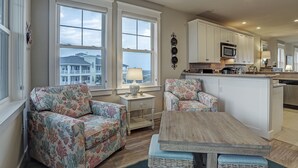 A spacious living area with two large windows, a kitchen with white cabinets, and a central island. - Beachfront at Pointe West. Walk to the beach and the club all just steps away! (Galveston)