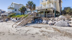 Una propiedad frente a la playa con un edificio de varios niveles, una escalera que baja a la playa y grandes rocas al frente.