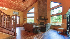 A spacious living room with a fireplace, wooden flooring, and large windows offering views of the outdoors.