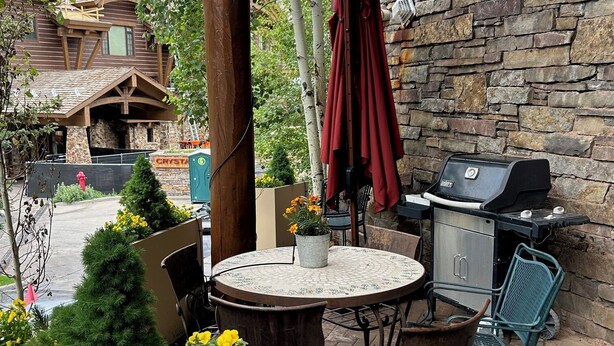 Une terrasse extérieure avec un mur en pierre, un barbecue, une table ronde avec des chaises et un pot de fleurs.