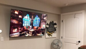 Un salon avec une cheminée, un grand téléviseur fixé au mur, un ventilateur et deux canapés en cuir.