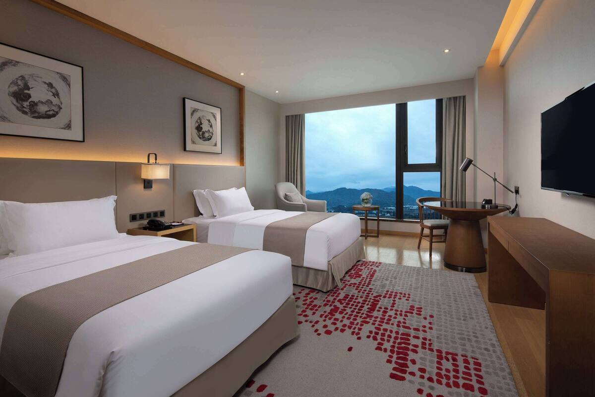 Habitación de hotel con dos camas, un escritorio, una silla, un televisor y un ventanal con vistas.