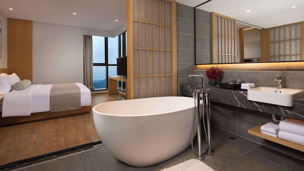 Une chambre d’hôtel moderne dotée d’une grande baignoire, d’un lit, d’une coiffeuse avec un miroir et offrant une vue sur l’extérieur.