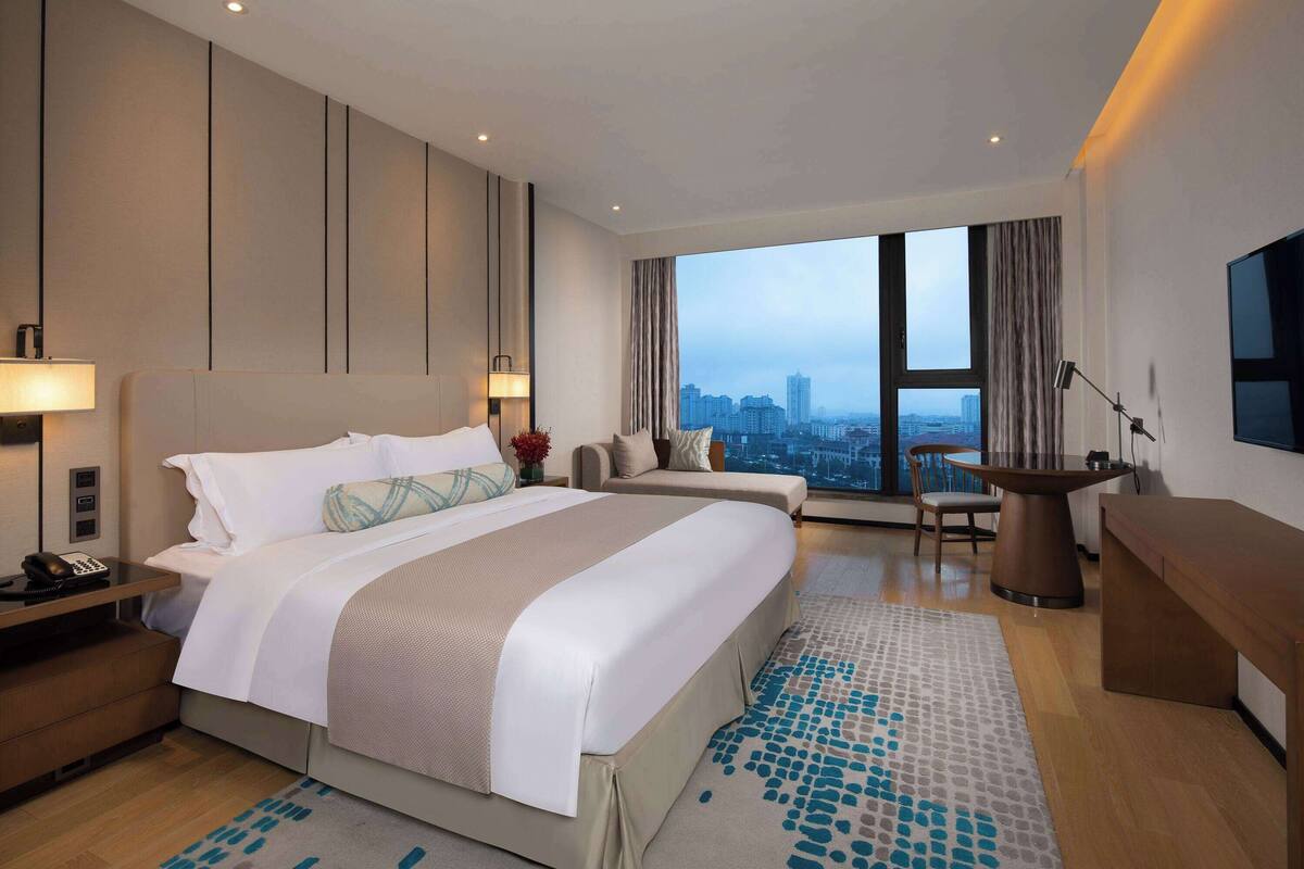 Una habitación de hotel moderna con una cama grande, un escritorio, una silla y vistas de la ciudad a través de amplias ventanas.