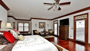 Une chambre avec un grand lit, un ventilateur de plafond, un plancher en bois, une télévision et une fenêtre avec des stores.