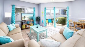 Un salon avec un canapé, une table basse et un téléviseur. Il y a un balcon d’où l’on peut admirer des immeubles et un palmier.