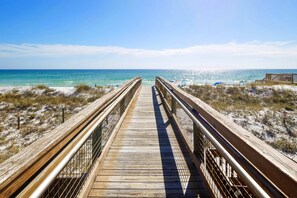 Ein hölzerner Steg führt zu einem Strand mit blauem Himmel und ruhigem Wasser.