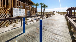 Une promenade en bois mène à une zone de plage où se trouvent un panneau d’information, des palmiers et une structure en bois.