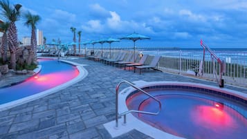 Un espace piscine avec des chaises longues et des parasols, une piscine éclairée en rouge et une vue sur l’océan.